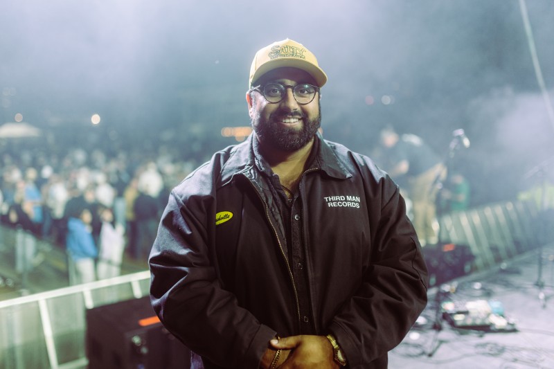 [small] Amit Mehta portrait: man with beard in black jacket, glasses and baseball cap with a festival crowd in the background